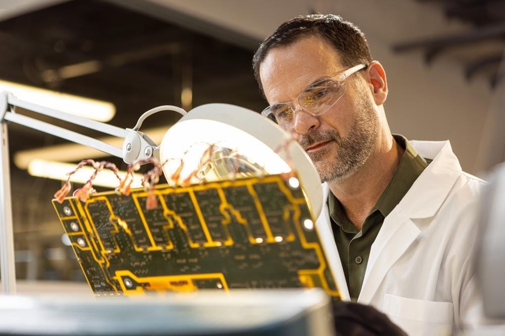 Un ingénieur en blouse de laboratoire et lunettes de protection inspecte un circuit imprimé sous une lampe loupe.