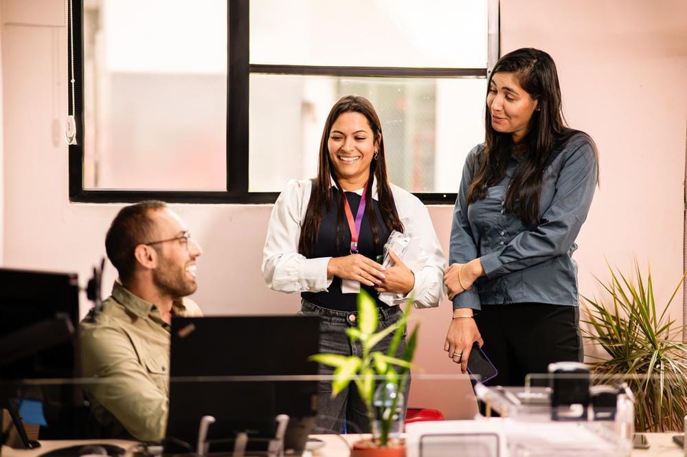 Trois collègues partagent un moment de rire autour d’un bureau, dans un espace lumineux et végétalisé.