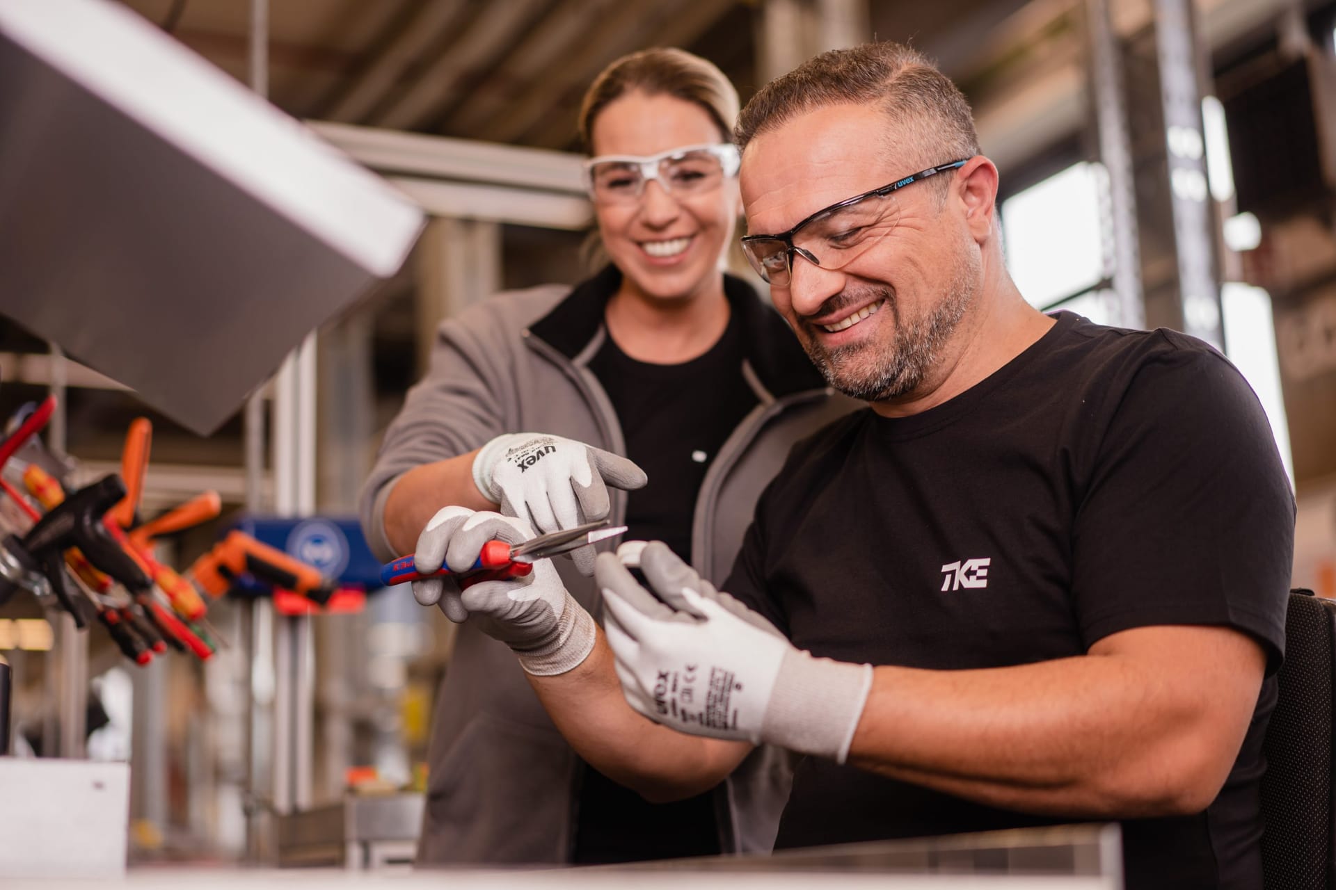 Deux collègues souriants, en lunettes de protection et gants de travail, manipulent ensemble un outil à l’établi.