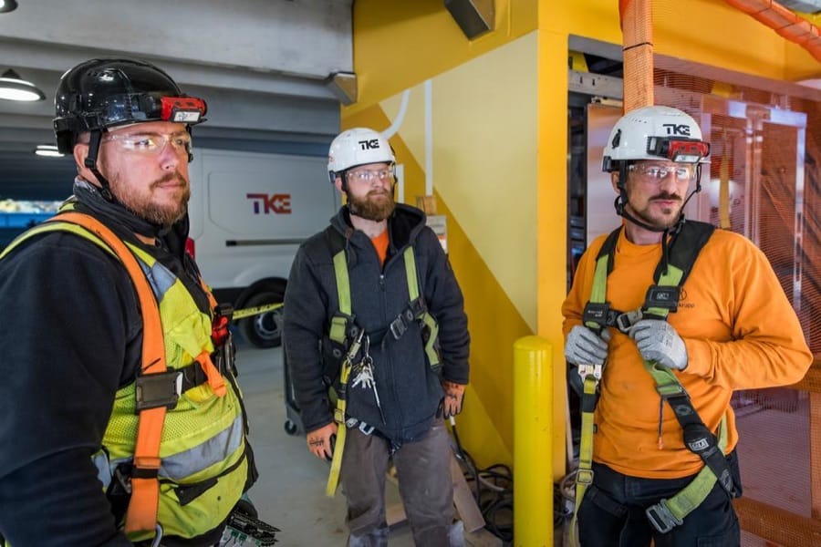 Trois techniciens TKE équipés de casques, de lunettes de protection et de harnais sur un chantier.